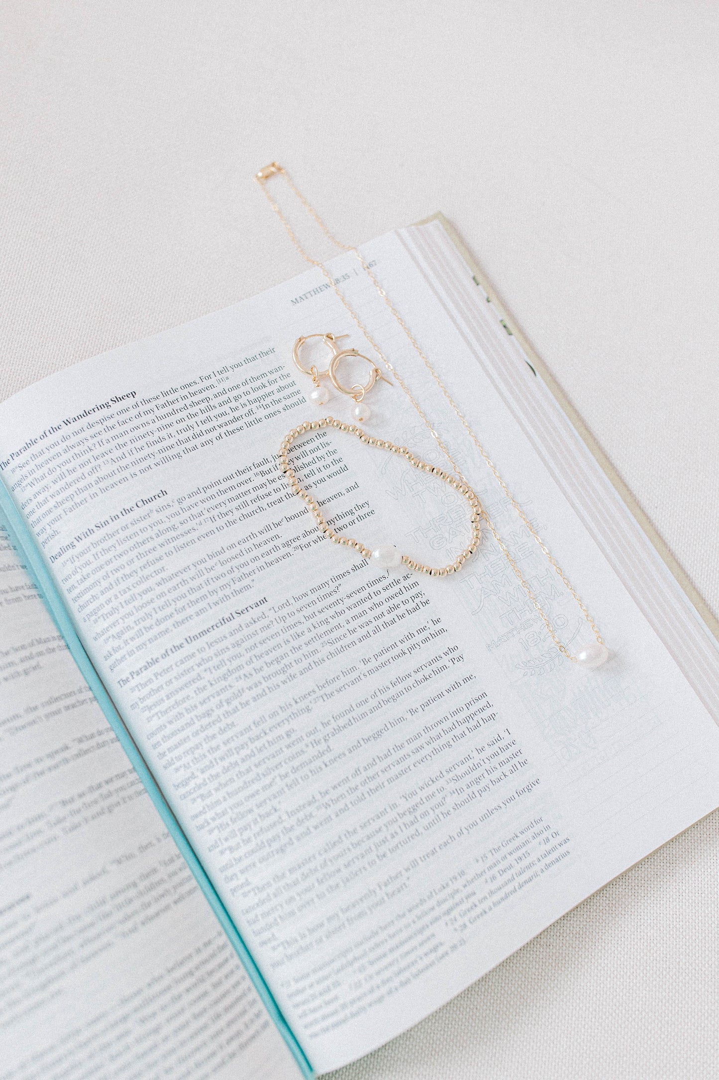 A photo of a bible with the He Left the 99 jewelry collection laying on top including a necklace, earrings, and bracelet. All gold with a single pearl.