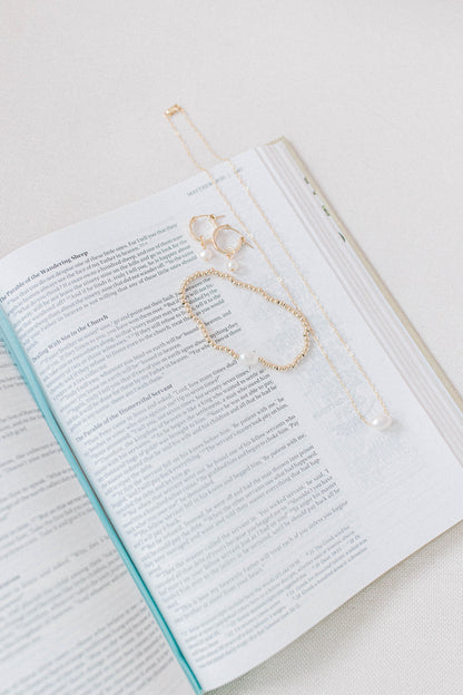 A photo of a bible with the He Left the 99 jewelry collection laying on top including a necklace, earrings, and bracelet. All gold with a single pearl.